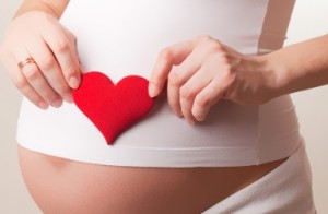 Photo of a pregnant belly with woman holding a red heart over it